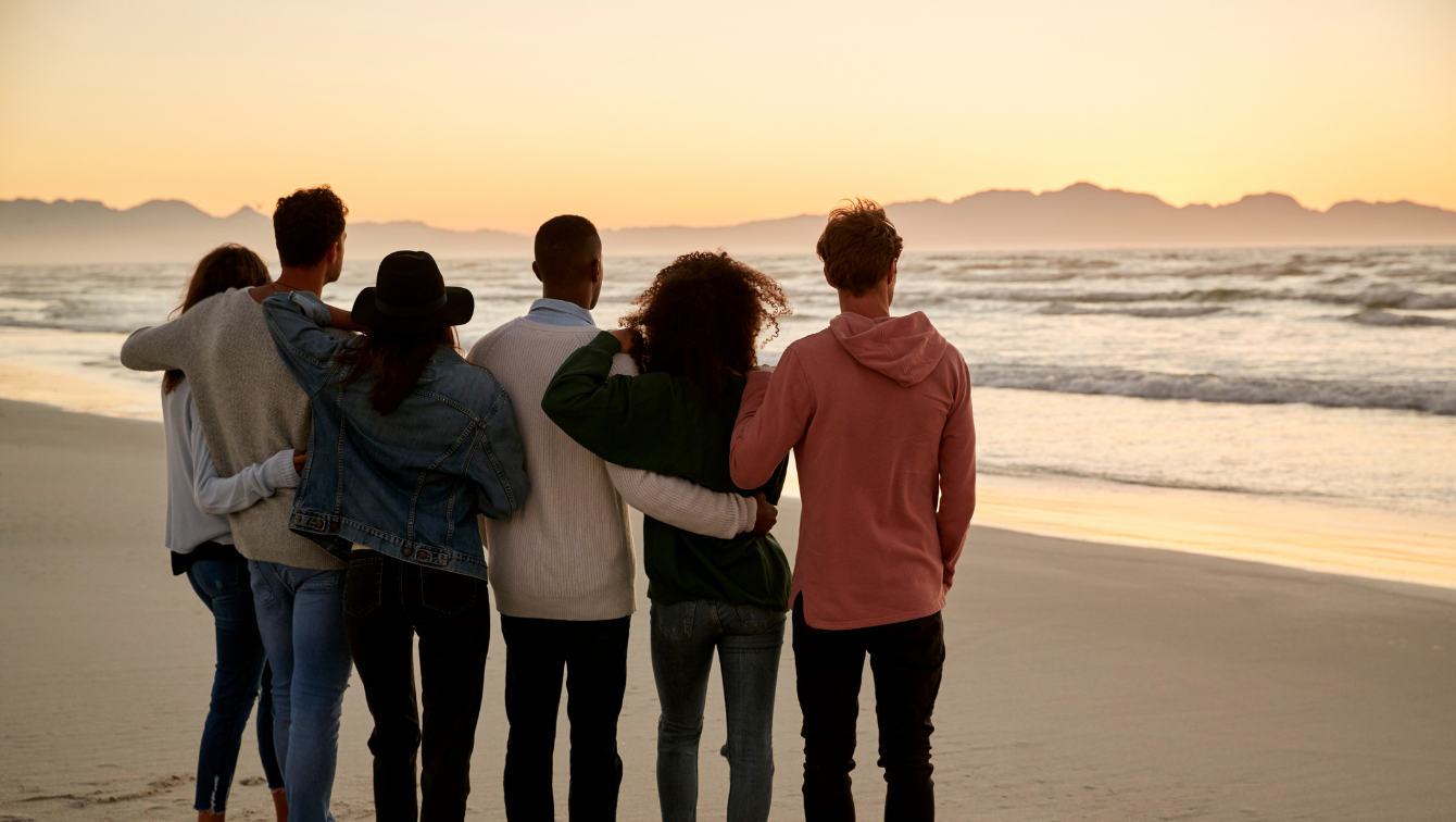 Six friends are standing side-by-side in a group hug on a beach, watching the sun rise