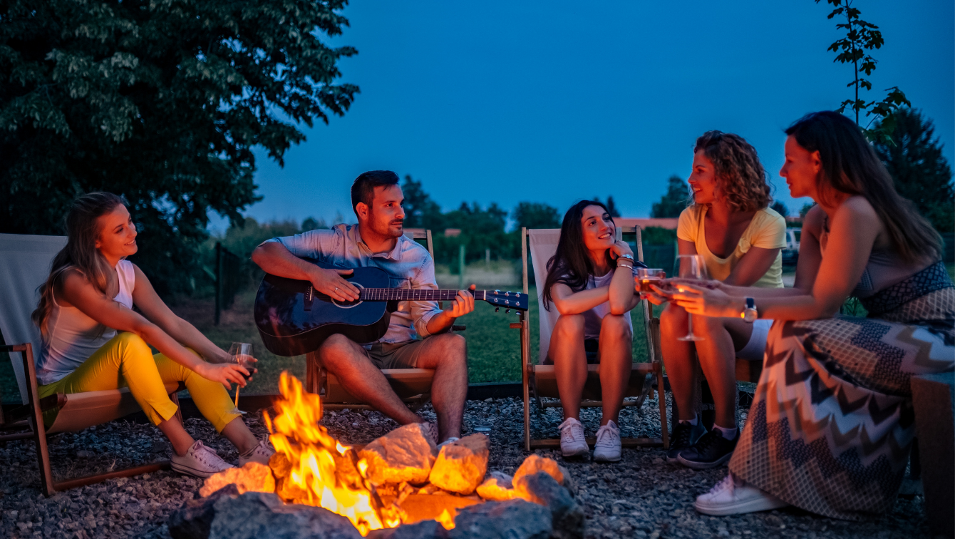 A group of friends are sitting around a campfire, some are having drinks, while one has stopped playing their guitar to listen to another peron talk