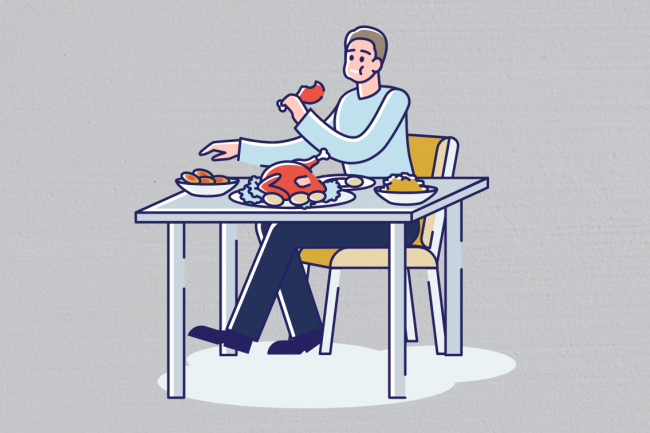 A man sitting on a table full of several foods, and eating a chicken leg. 