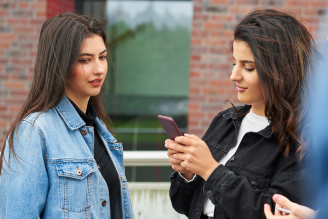 Two friends chat. One is fiddling with her phone, while the other looks at the device and seems uncomfortable. 