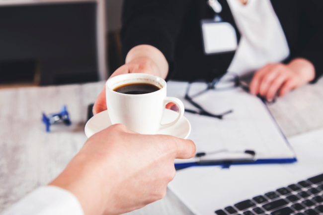 One person offers a cup of coffee to another one over a work desk. Only their hands show in the image. 