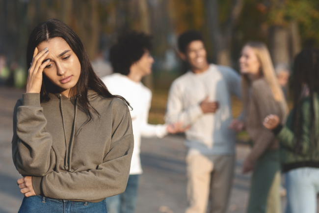 A teen seems sad and left out while a group of friends is chatting happily in the background (at a distance). 