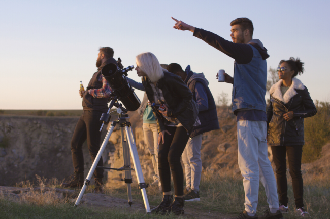 A group of friends at a mountain top with a telescope. One points to the sky, and another looks through the telescope. 