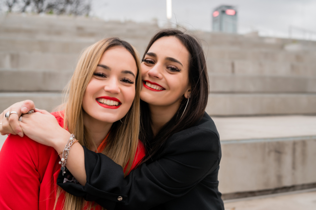 Two friends hug each other side by side. They're both smiling. 