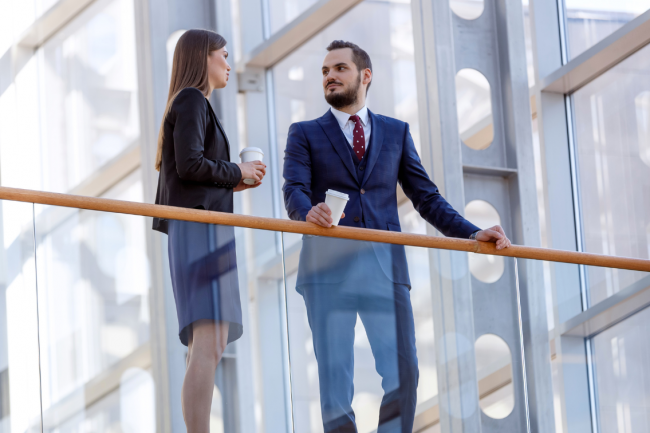 Two people drink coffee and talk to each other in the external area of an office building. 