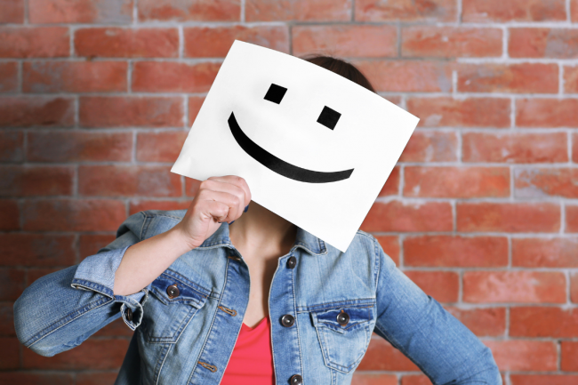 A person holds a sheet of paper over their face. The sheet has a smiling icon printed on it. 