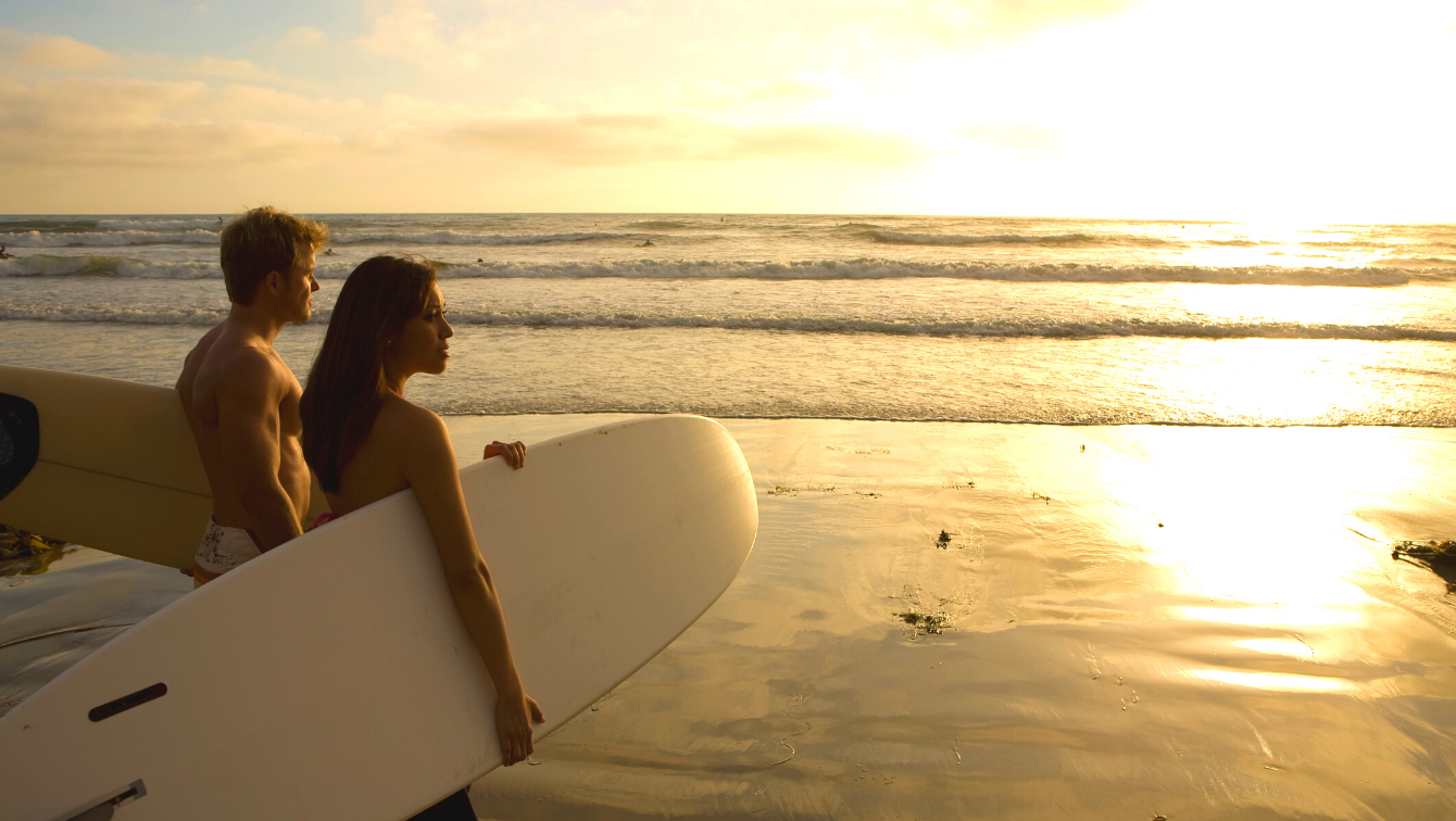 A couple watching the sunset by the beach while holding surf boards. 