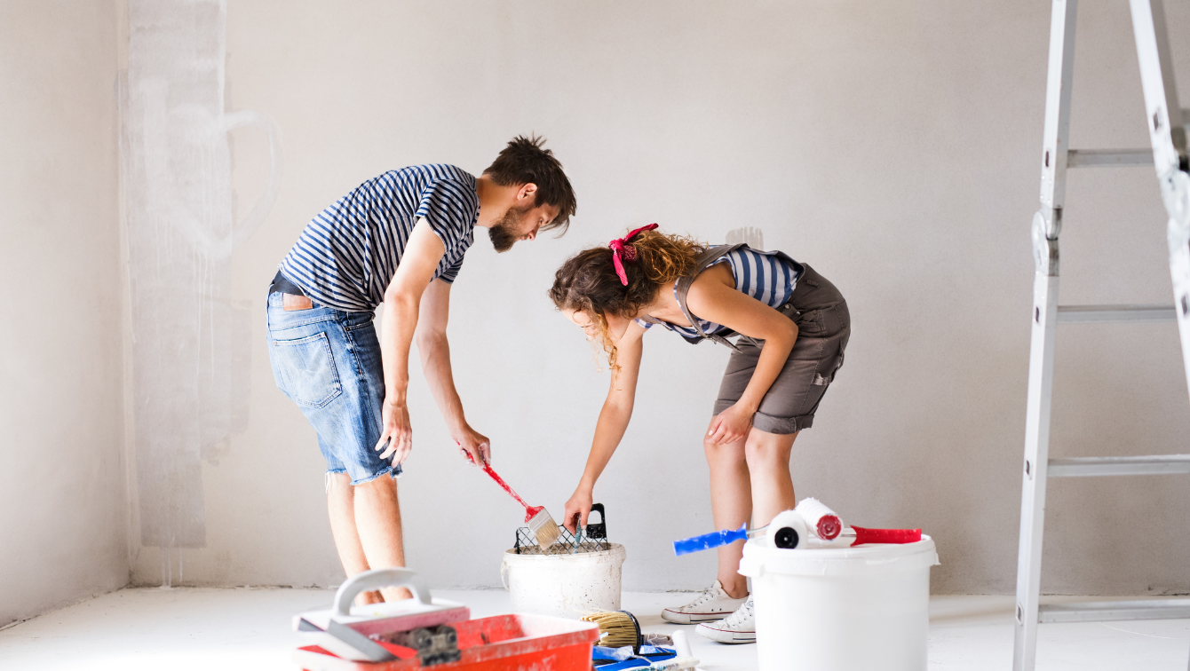 A couple painting a wall together in a empty room. There are some painting instruments around them. 